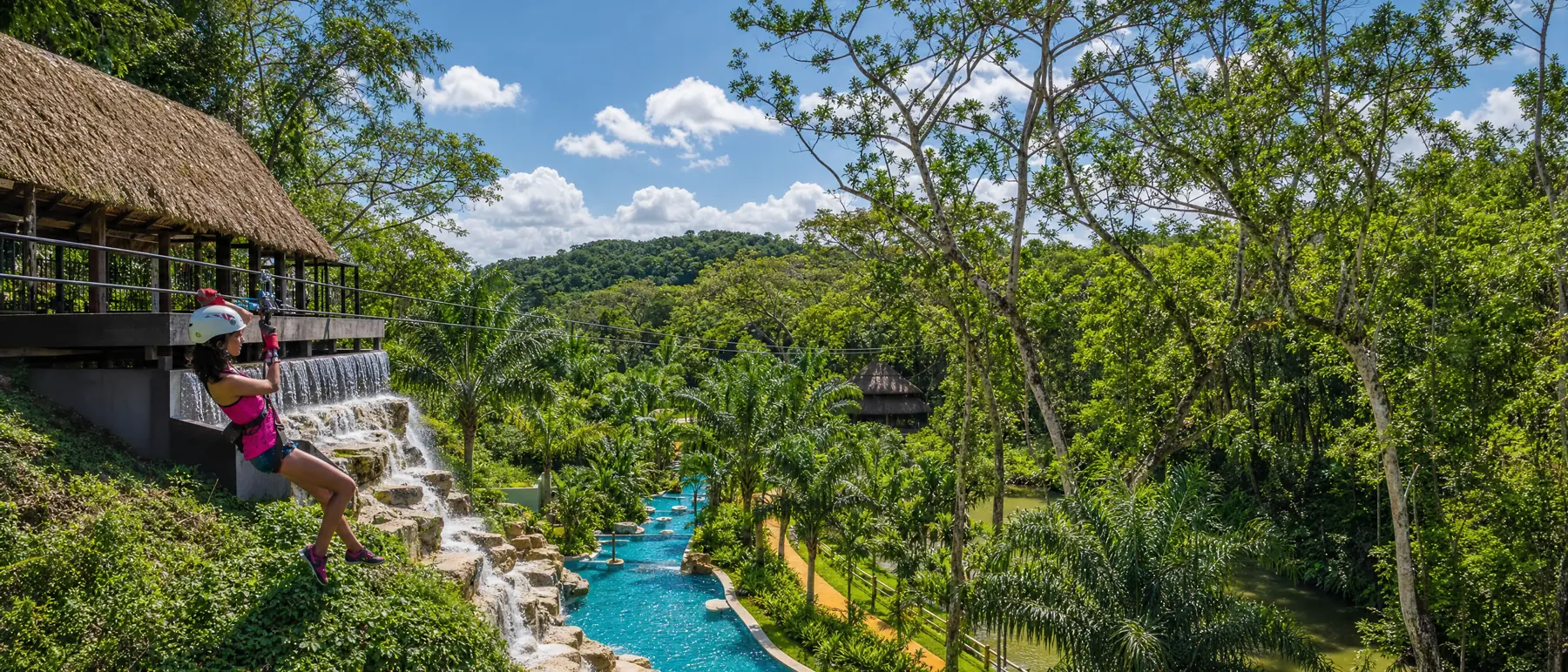 A zipline rider setting off from a thatched platform above a waterfall and a winding blue water channel, palms and forested hills beyond