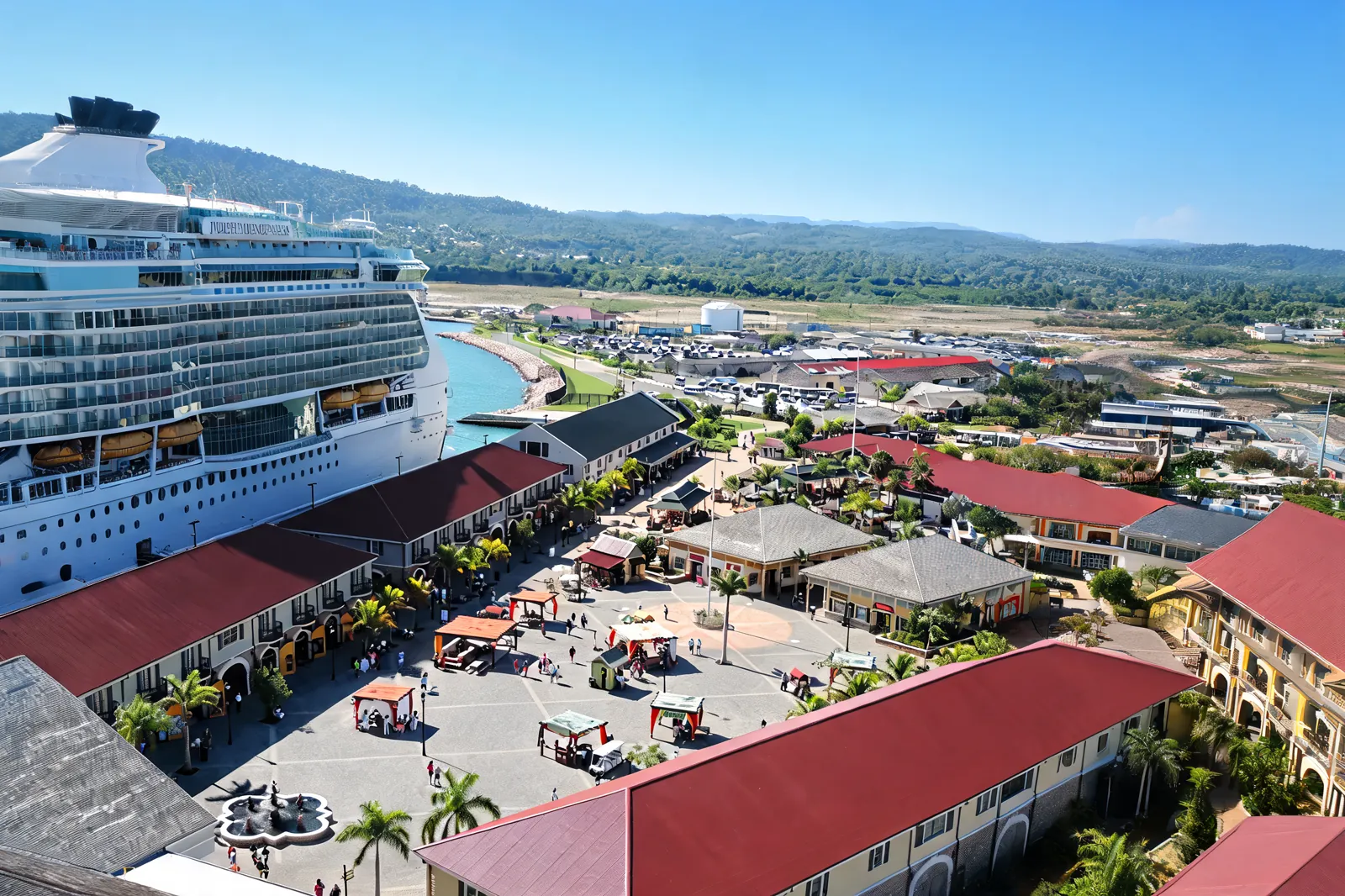 An aerial view of Falmouth's cruise port, a docked ship beside red-roofed terminal buildings, palms and a plaza, green hills behind