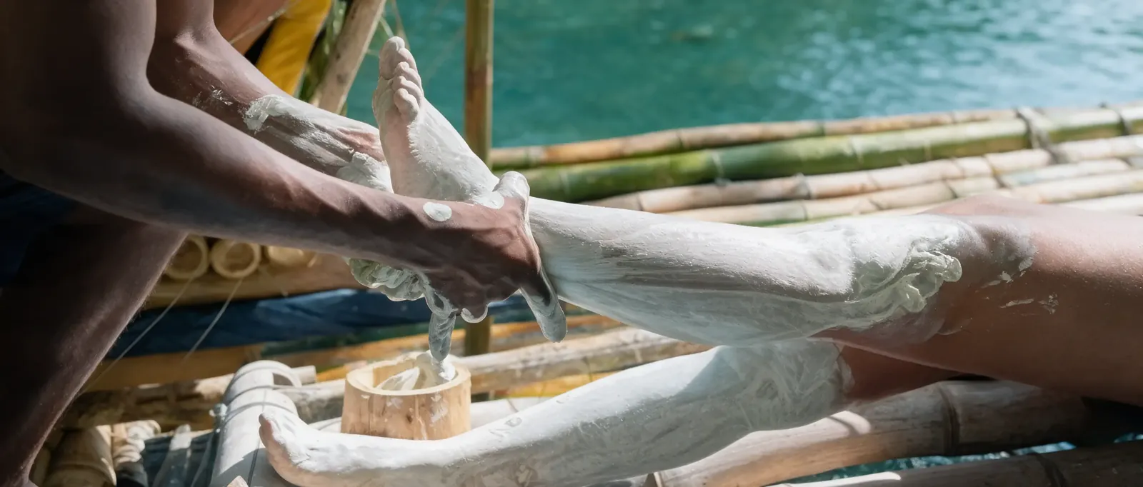 A raft captain smoothing white limestone paste onto a guest's leg on a bamboo raft, green river water behind