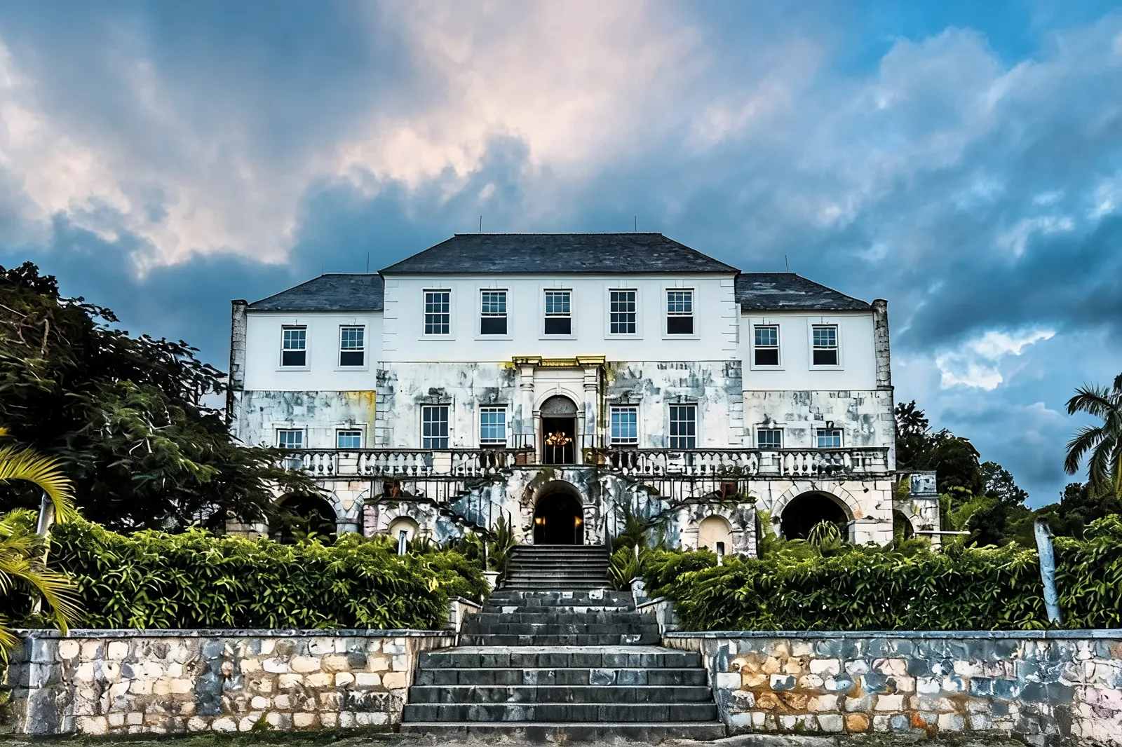 The Rose Hall great house head-on from the foot of its stone staircase, white Georgian facade under a dark cloudy sky