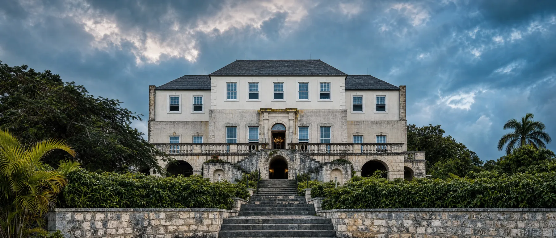 The Rose Hall great house head-on above its stone terrace and staircase, white Georgian facade framed by palms under a heavy grey sky
